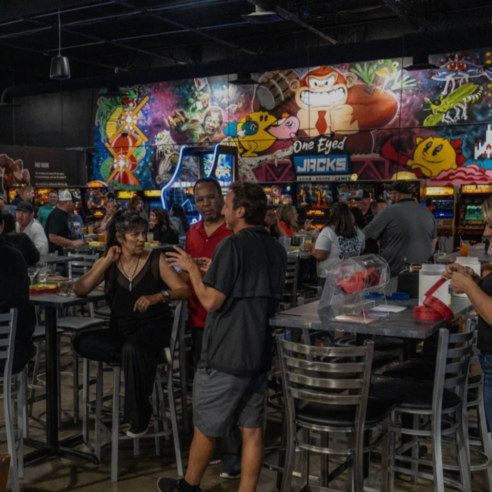 Patrons socialize at high-top tables in The Pickled Goat Bar & Grill, with arcade game murals in the background.