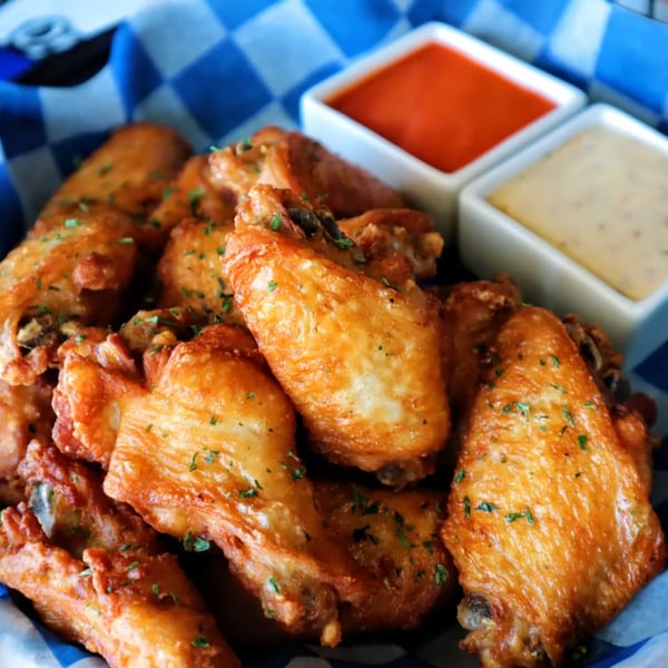 Crispy chicken wings with parsley flakes, served in a blue checkered basket with two dipping sauces.