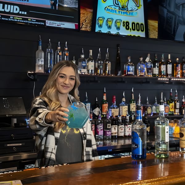 A smiling bartender at The Pickled Goat Bar & Grill holds out a blue cocktail.