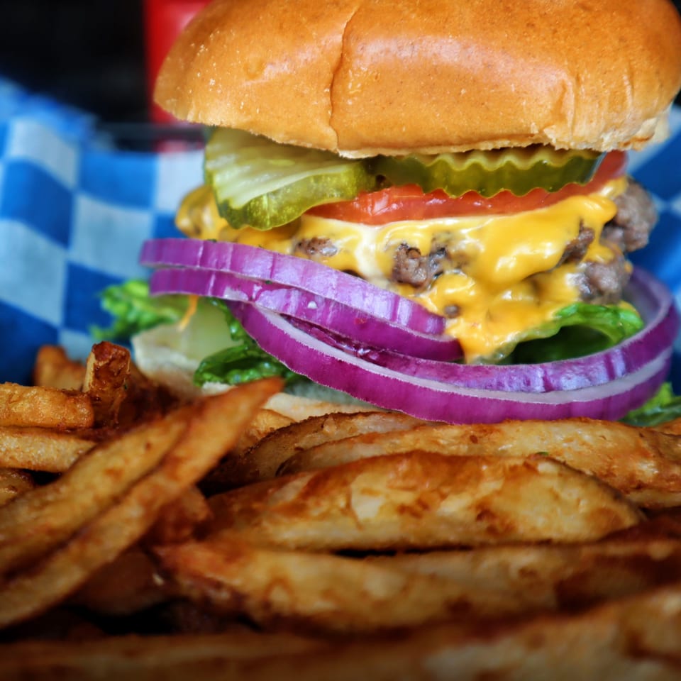 A double cheeseburger with lettuce, red onion, tomato, pickles, and fries in a blue checkered basket.