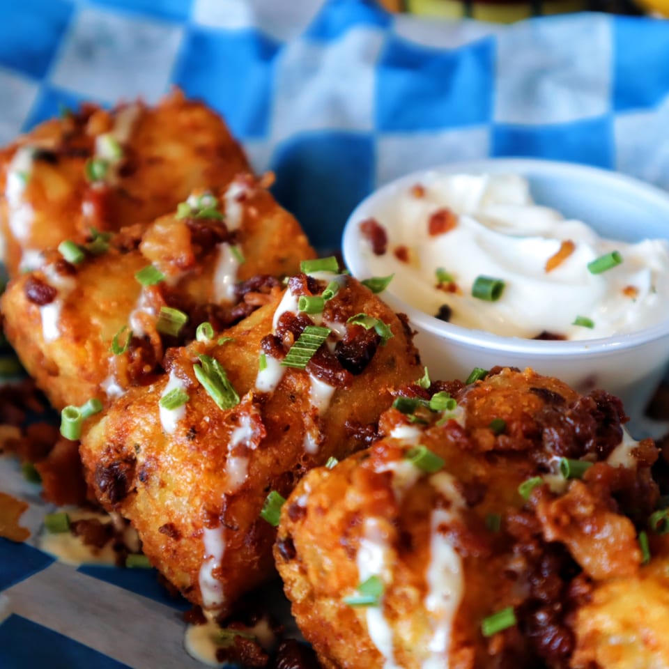 Golden-brown tater tots topped with bacon, white sauce, and green onions, with a side of dipping sauce.