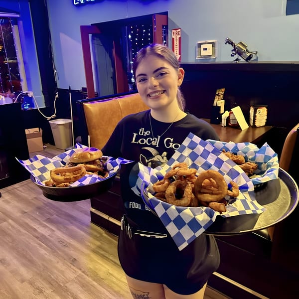Smiling server at The Pickled Goat Bar & Grill holding trays of burgers, onion rings, and chicken.