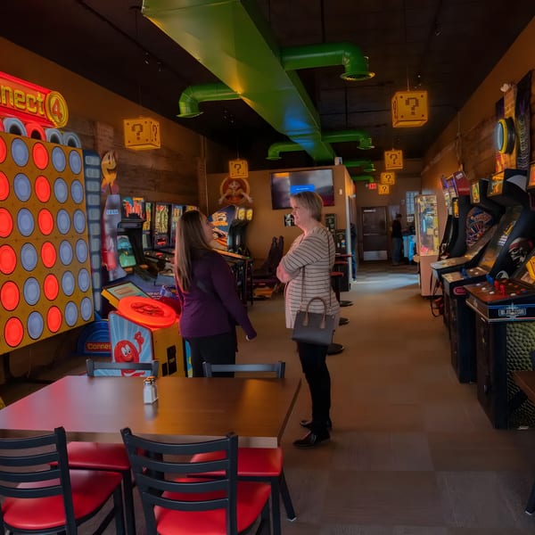 Two women talking in a dimly lit arcade room with various games and Mario-themed decor.
