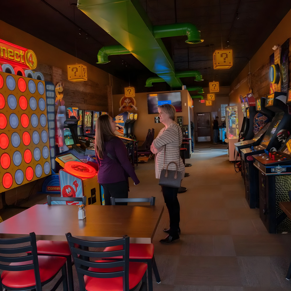 Two women talking in a dimly lit arcade room with various games and Mario-themed decor.