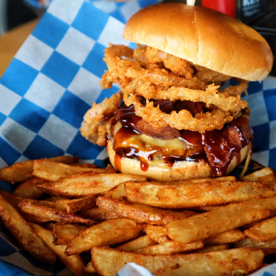 A Pickled Goat Bar & Grill burger with onion rings, bacon, cheese, and BBQ sauce, served with fries.