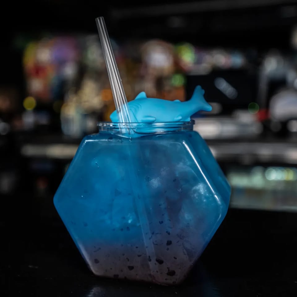 A blue cocktail in a hexagonal glass, topped with a shark lid and straw, on a dark bar.