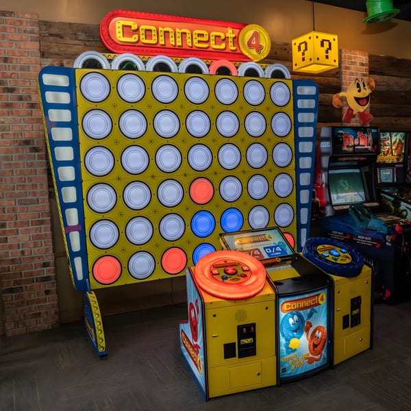 A giant Connect 4 arcade game with red and blue pieces, alongside other arcade machines and Pac-Man decor.