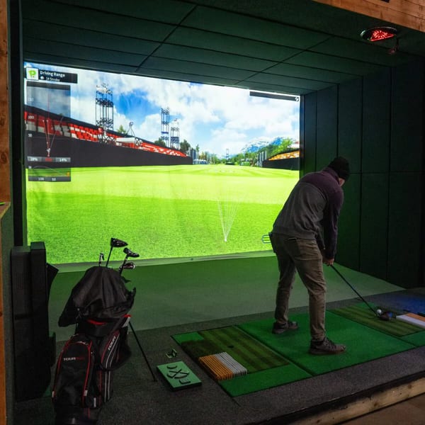 A man in a dark beanie and jacket plays golf on a simulator with a large screen showing a driving range.
