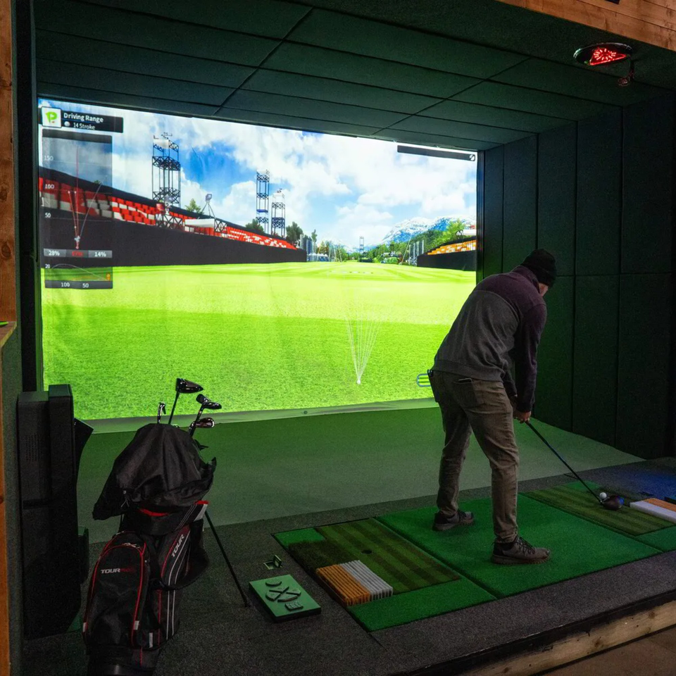 A man in a dark beanie and jacket plays golf on a simulator with a large screen showing a driving range.