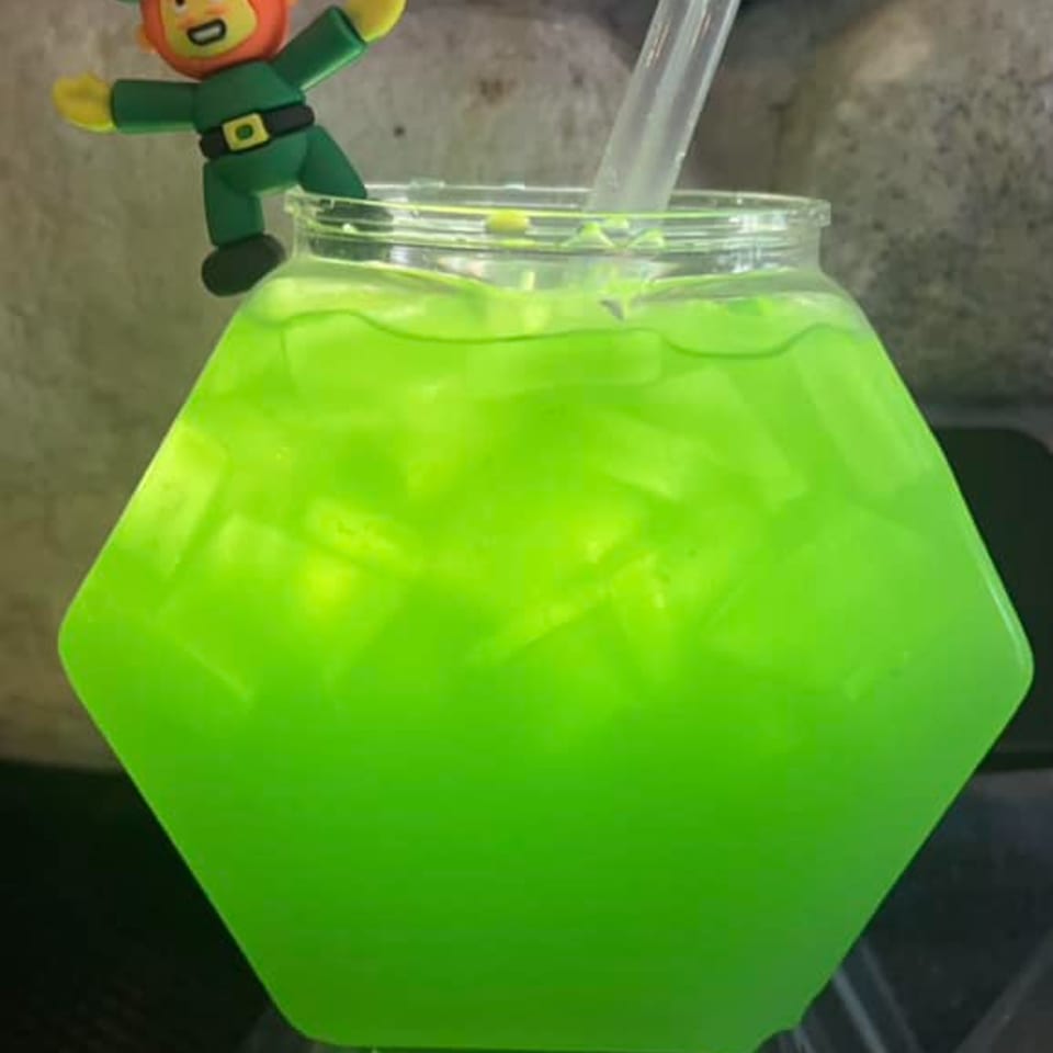 A bright green drink in a hexagonal glass with a leprechaun figurine clinging to the rim.