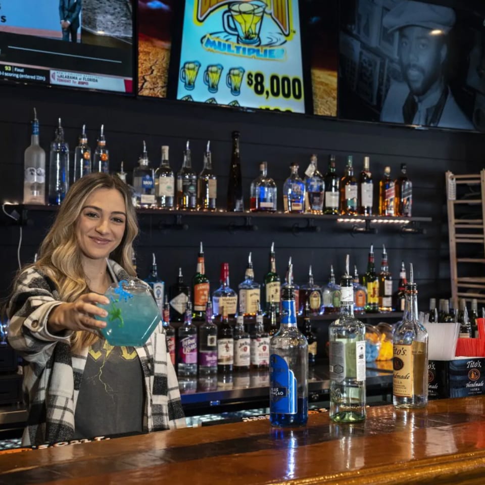 A smiling bartender at The Pickled Goat Bar & Grill holds out a blue cocktail.