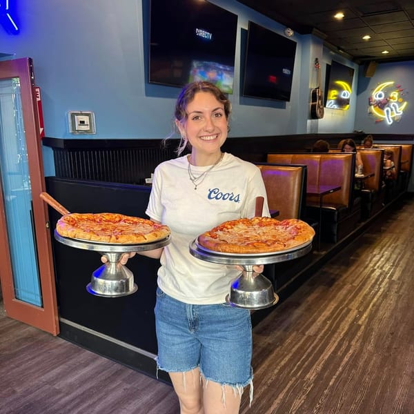 Smiling server holding two pizzas on stands in The Pickled Goat Bar & Grill.