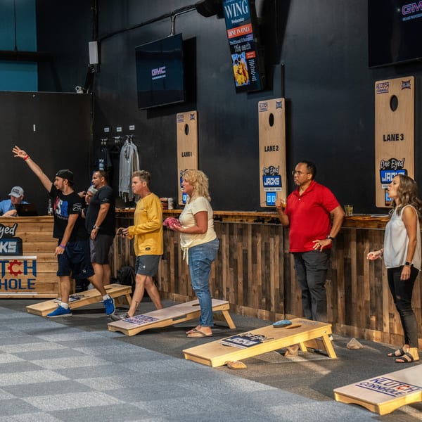 People playing cornhole at The Pickled Goat Bar & Grill, with scoreboards and a wing special visible.