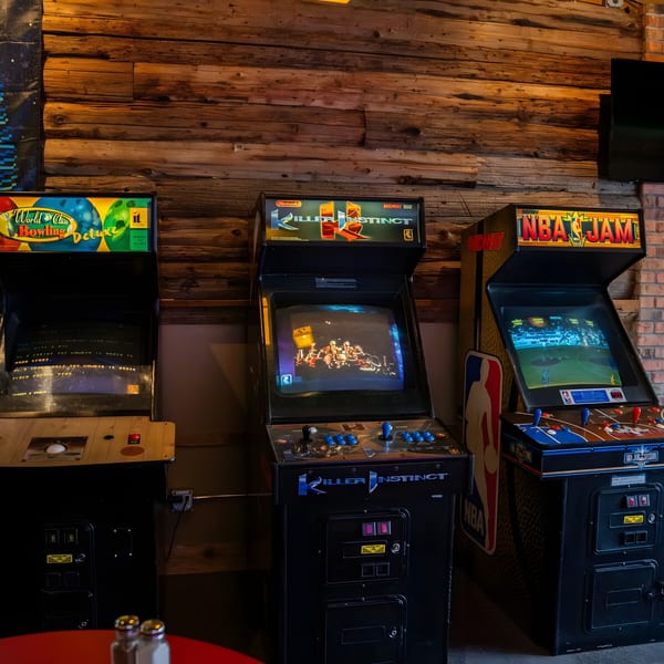 Three vintage arcade games, including Killer Instinct and NBA Jam, stand against a rustic wooden wall.