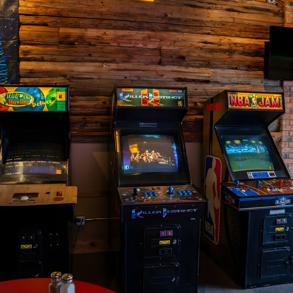 Three vintage arcade games, including Killer Instinct and NBA Jam, stand against a rustic wooden wall.