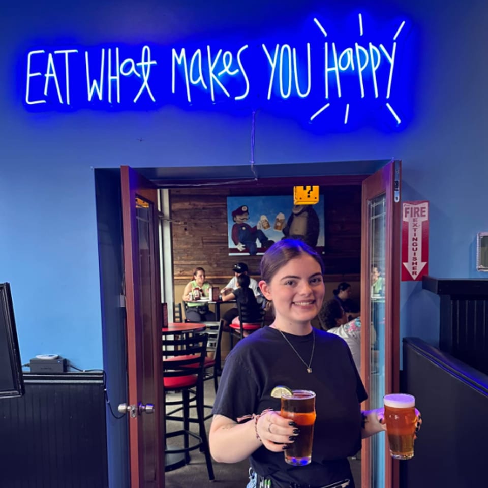 A smiling server holds two beers in front of a blue neon sign that reads "EAT WHAT MAKES YOU HAPPY".